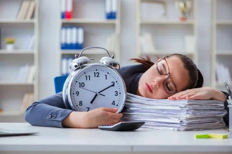 women sleeping at desk ontop of her papers while holding a clock