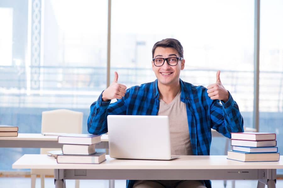 male student sitting at desk with laptop open and holding both thumbs up