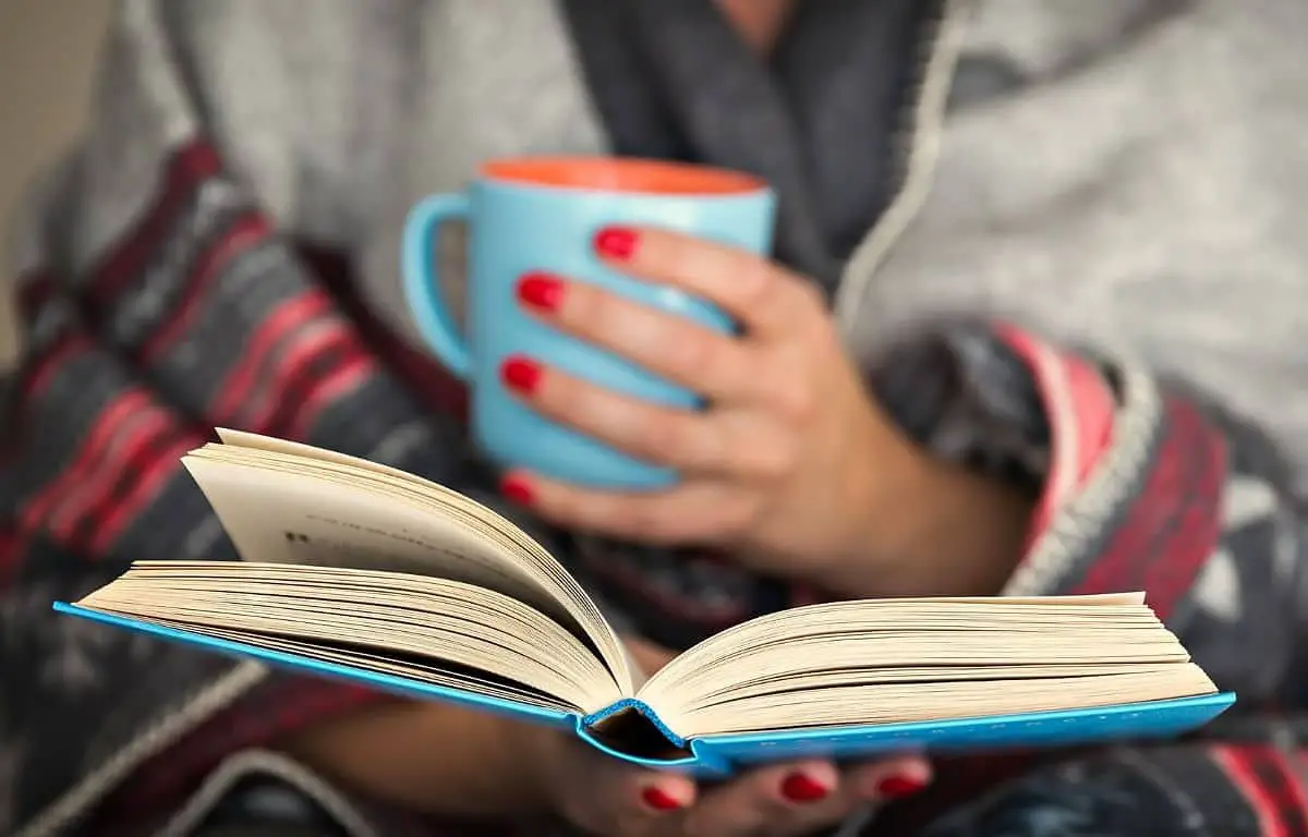 female hand holding a cup of tea in her left hand and rereading a book by holding it in her right hand